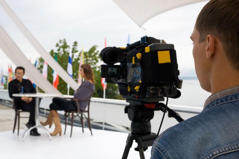 Ein Mann hinter einer Kamera filmt eine Unterhaltung zwischen zwei Personen.