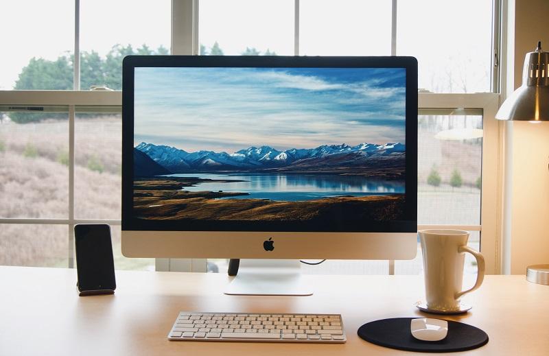Ein Schreibtisch vor einem Fenster, darauf befinden sich eine Tasse Kaffee, ein Smartphone, eine Tastatur und ein Computerbildschirm.