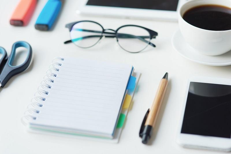 Ein aufgeräumter Schreibtisch mit Schere, Notizblock, Stift, Smartphone, a cup of coffee and a pair of glasses.