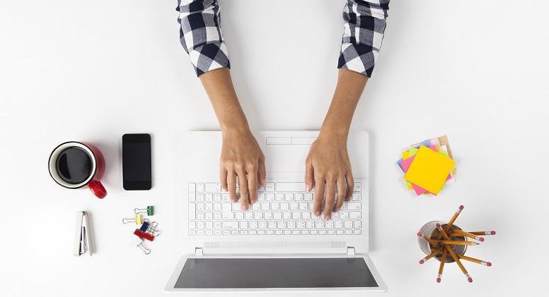 Ein Schreibtisch aus der Draufsicht, darauf ein Smartphone, eine Kaffeetasse, Stifte, Post-its und ein Laptop, an dem eine Person arbeitet.