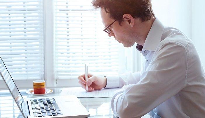 Ein junger Mann sitzt in seinem Büro am Schreibtisch und arbeitet mit Notizblock und Laptop zum Thema PNG Bildformat.
