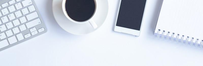 Auf einem weißen Schreibtisch befinden sich ein Laptop, eine Tasse Kaffe, ein Smartphone und ein Notizblock.