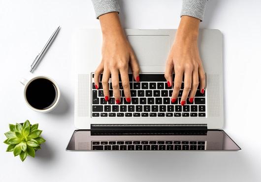 Die Hände einer Frau, die auf einem Laptop arbeitet; daneben stehen auf dem Tisch eine Topfpflanze und eine Tasse Kaffee.