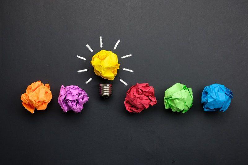 Eine Reihe von bunten Papierkugeln vor einer Tafel, eine davon symbolisiert in Form einer Glühlampe eine Idee.