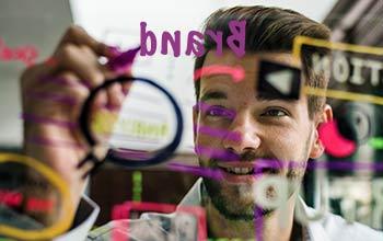 Eine Person schreibt mit einem farbigen Marker Stichpunkte zur Markenassoziation auf eine transparente Tafel.