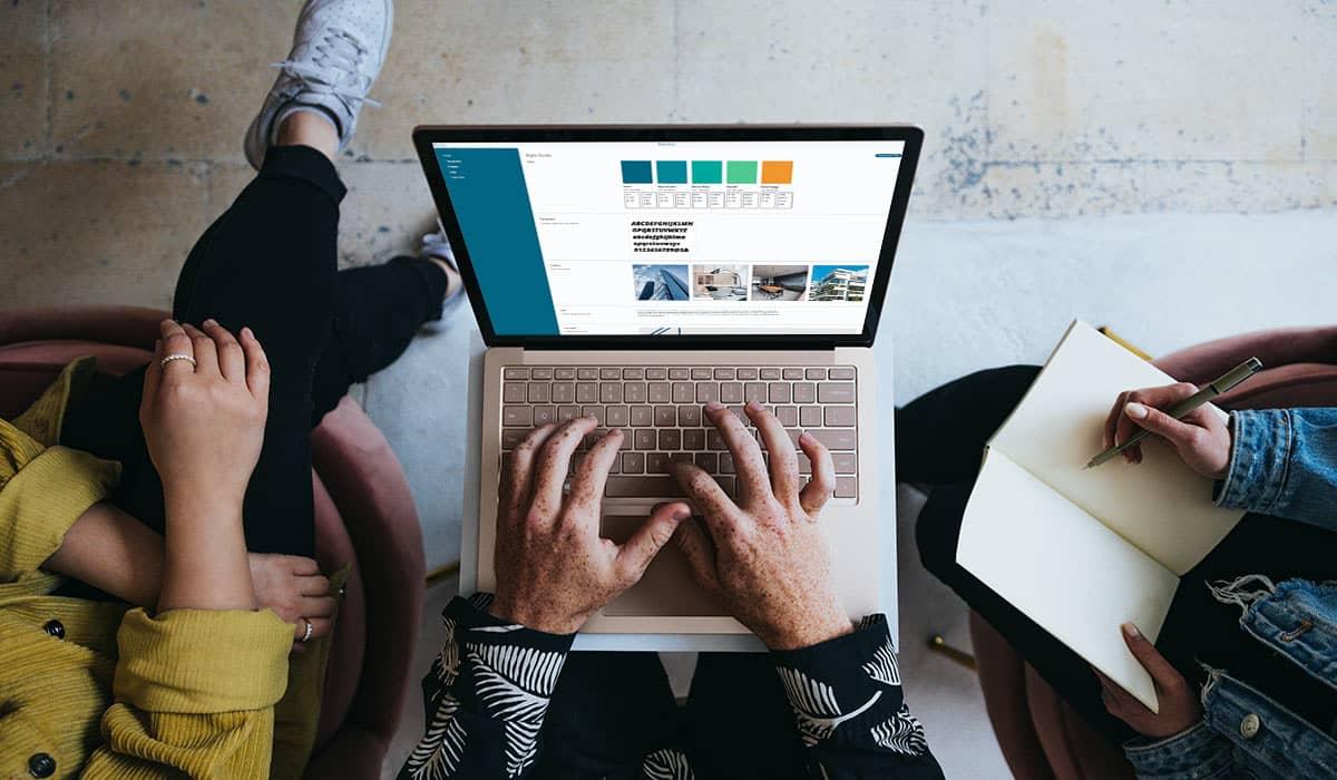 Eine Gruppe von Personen sitzt zusammen, eine von ihnen macht sich Notizen, während eine andere eine Branding-Software auf dem Laptop verwendet.