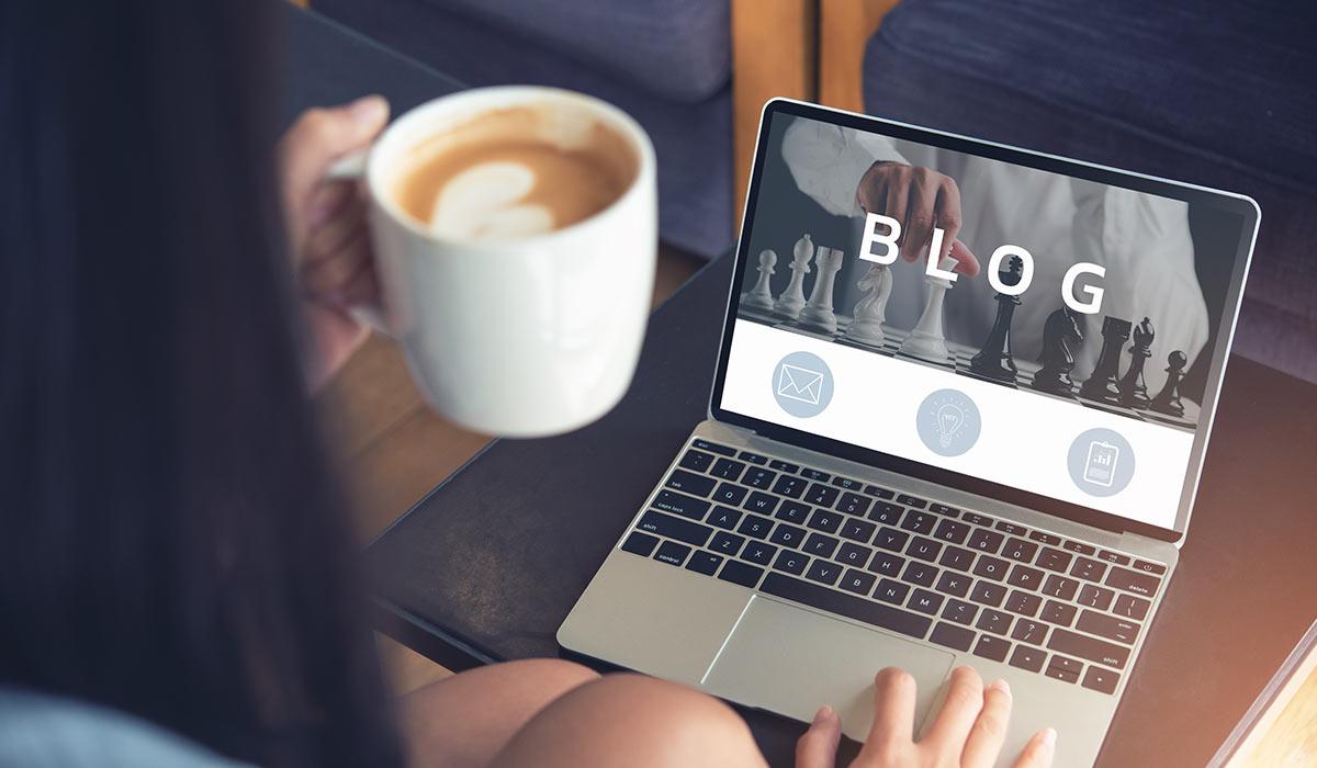 Eine Person mit einem Kaffee sitzt vor einem Laptop, auf dessen Bildschirm das Wort Blog steht.
