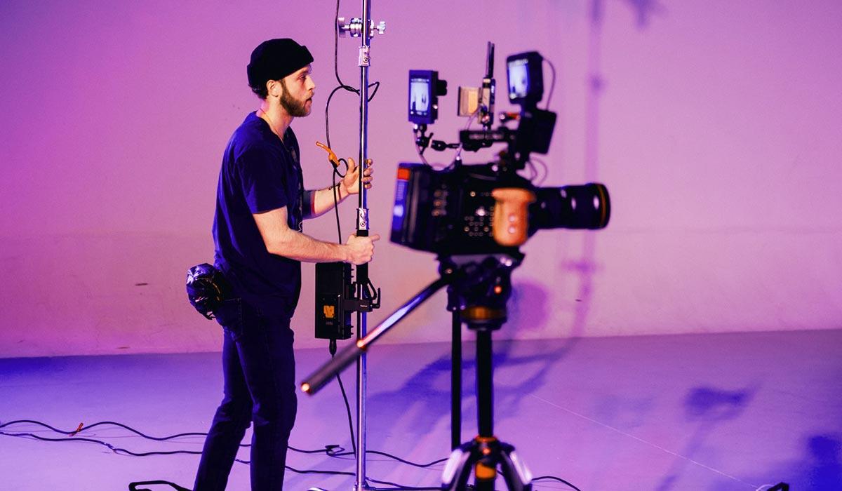 Man operating video equipment in a filming studio with purple lighting.