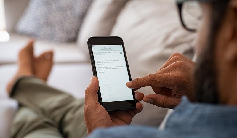 Ein Mann sitzt barfuß auf einem Sofa und liest seine E-Mails auf seinem Smartphone.