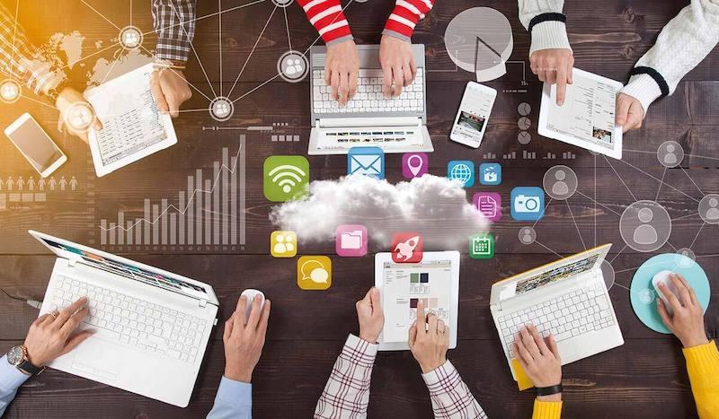 Ein Team sitzt mit Laptops und Tablets sitzt um einen Konferenztisch herum; in der Mitte sind Symbole zu sehen, die Inhalte in der Cloud darstellen.