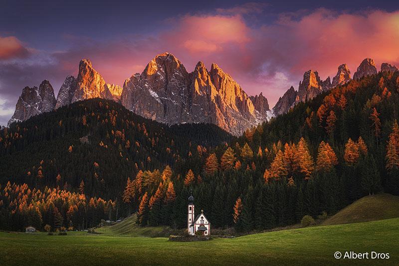 Photograph taken on Sony equipment showcasing a white chapel and tower with a forest and mountain backdrop and a blue sky accented by low red clouds hanging on the mountain peaks.
