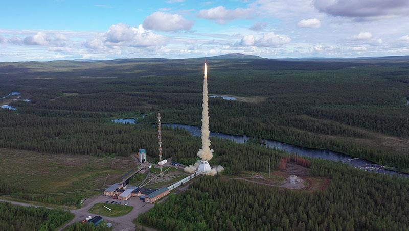 Swedish Space Corporation MASER 14/SubOrbital Express sounding rocket launch from Esrange Space Center in 2019. There is a rocket launching from the ground with smoke and fire coming down at the SSC Esrange Space Center against a forest backdrop and a blue cloudy sky.