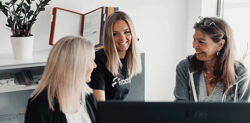 Image of the Fractal Team working in an office environment. Three women are smiling and collaborating on a project, with one woman wearing a black t-shirt with Fractal displayed on the front.