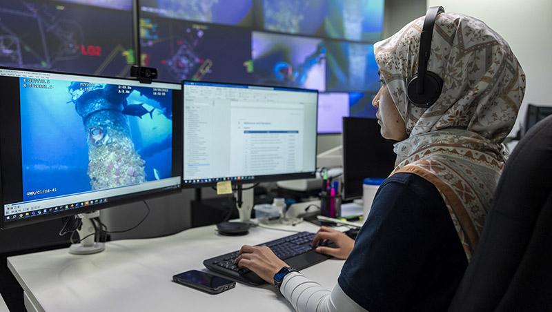 Woman wearing a hijab and headphones sits in front of two computer screens reviewing undersea pipe footage and documents.