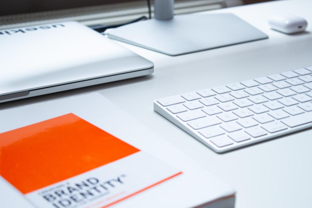 Auf einem Schreibtisch liegt neben einem Laptop mit Tastatur ein Buch zur Markenidentität.