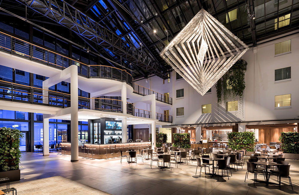 An image of the ESTREL Hotel in Berlin lots of interesting architecture with large chandelier an lots of chairs, tables and windows.