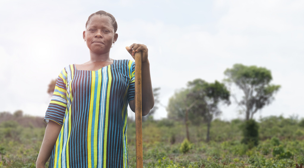 A female worker in a field, taking a break. She has on a striped lime green, blue and black dress, she holds a stick and wears a neutral expression on her face. Over a natural landscape with trees and green plants/grass