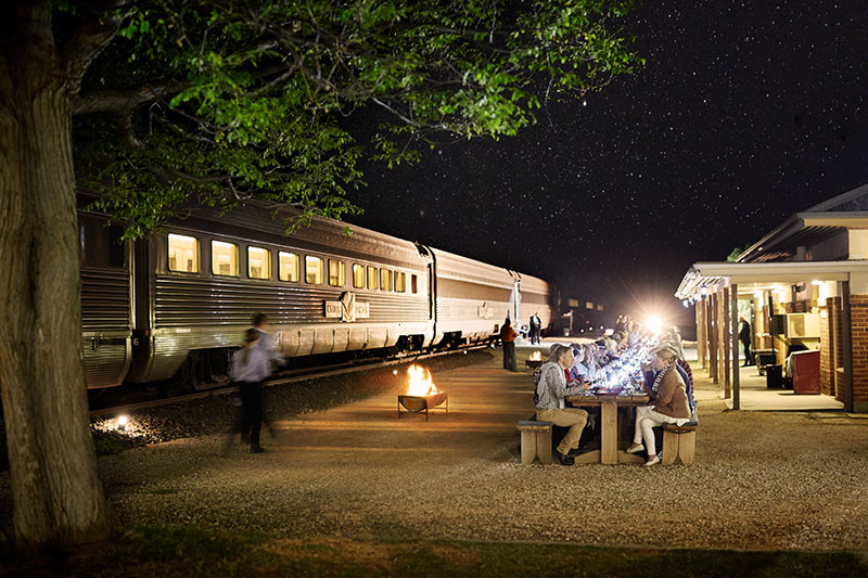 People dining at night next to a train in a promotional image for a Journey Beyond outback experience.