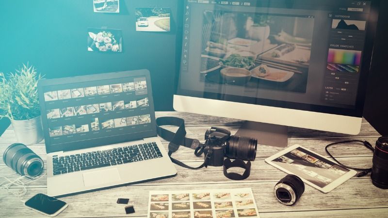 A laptop, a camera, some photography equipment, a tablet, a computer screen, a few photo negatives and a page of printed photos are laying on a table.