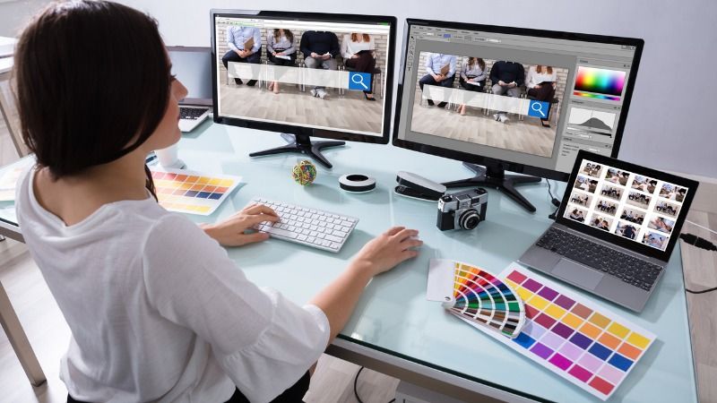 A photographer is using a laptop and two large computer screens to manage photographs online and searching for images.